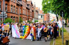 Eine große Gruppe von Menschen läuft auf der Straße. Sie halten einen Banner mit der Aufschrift "Pride Lüneburg". Im Hintergrund auf dem Banner ist die erweiterte Pride-Flag (weiß, hellblau, pink, braun, schwarz, lila, blau, grün, gelb, orange, rot, gelb mit rotem Kreis) zu sehen. Menschen halten verschiedene Flaggen und Banner hoch. Die Stimmung wirkt motiviert.